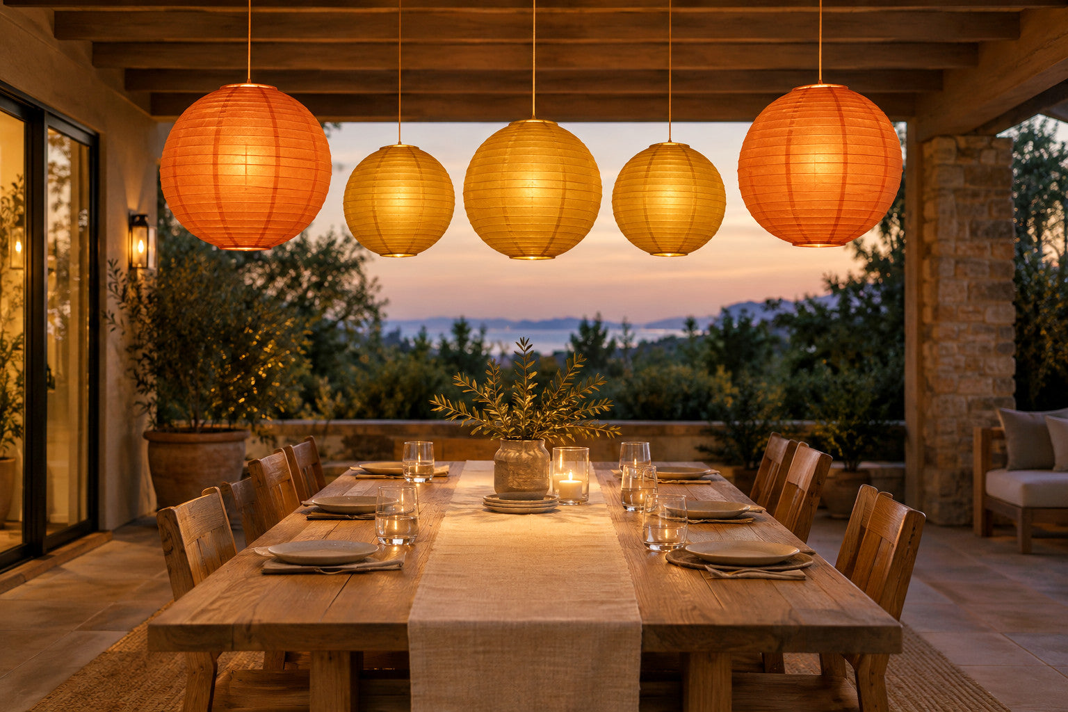 Five gold and orange paper lanterns with warm white lights hanging in a row above a set dining table on a covered patio.