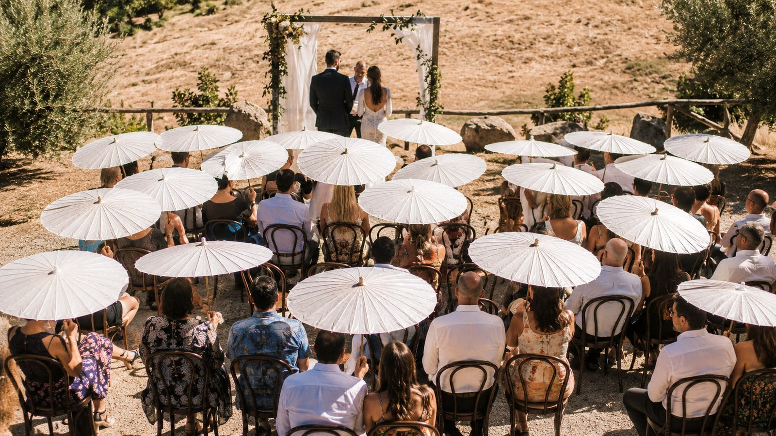Wedding guests seated in full sun at an outdoor ceremony, most holding open white parasols with long bamboo handles